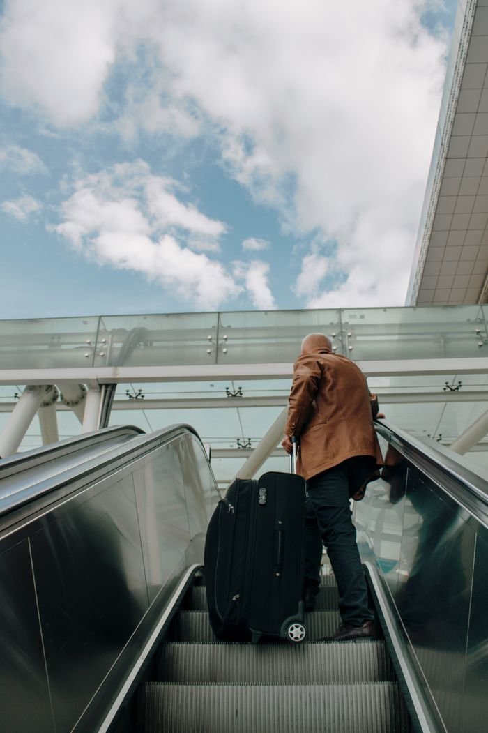 Home Adult male riding an escalator with luggage in an airport, traveling with bright sky overhead.